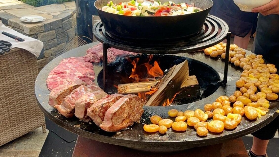 Ein Grill mit verschiedenen Fleischsorten und Gemüse wird zubereitet. Im Hintergrund sind grüne Hügel und ein klarer Himmel zu sehen. | © Tourismusverband Oststeiermark