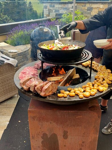 Egy grillezett különböző húsfélékből és zöldségekből álló étel készül. A háttérben zöld dombok és tiszta ég látható. | © Tourismusverband Oststeiermark
