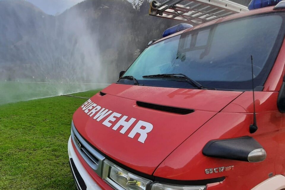 Ein rotes Feuerwehrfahrzeug steht auf einer grünen Wiese. Im Hintergrund sind Berge und ein Wassernebel zu sehen. | © FF Mößna - Manuel Wieser