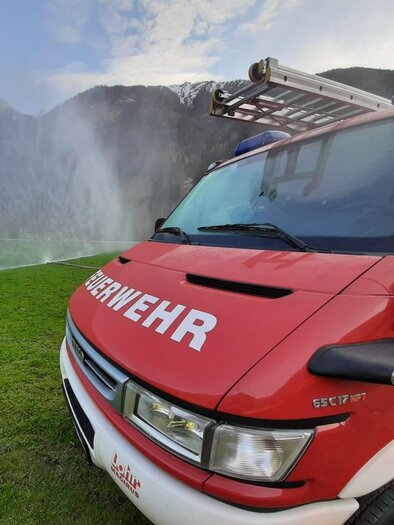 Ein rotes Feuerwehrfahrzeug steht auf einer grünen Wiese. Im Hintergrund sind Berge und ein Wassernebel zu sehen. | © FF Mößna - Manuel Wieser