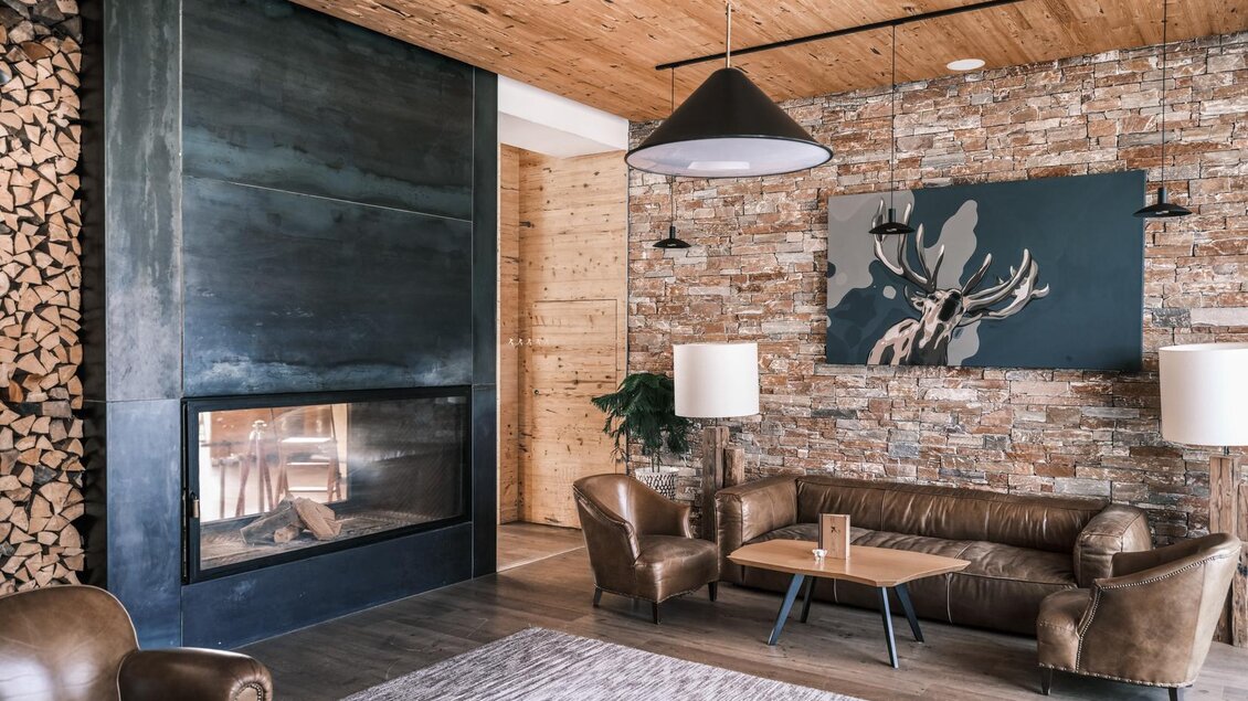 Ein modernes Wohnzimmer mit einem eleganten Kamin und Holzauflage an der Wand. Die Einrichtung besteht aus gemütlichen Ledermöbeln und stilvollen Lampen. | © Falkensteiner Hotels &amp; Residences