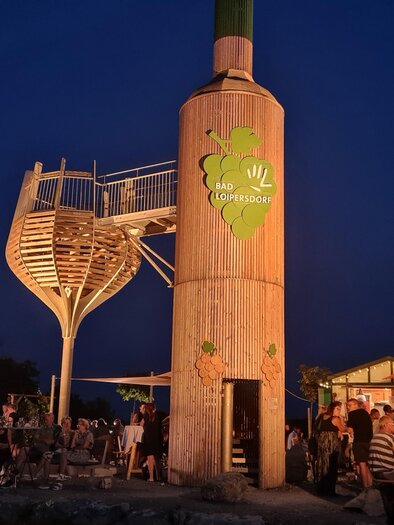 Eine belebte Freiluftbar mit einer einzigartigen Holzstruktur bei Nacht. Menschen sitzen drinnen und draußen und genießen die entspannte Atmosphäre. | © Gemeinde Bad Loipersdorf