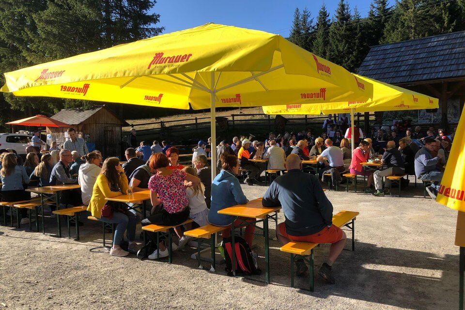 Eine belebte Außenterrasse mit vielen Gästen, die unter gelben Sonnenschirmen sitzen. Im Hintergrund sind Bäume und eine rustikale Hütte zu sehen. | © Johannes Lintschinger