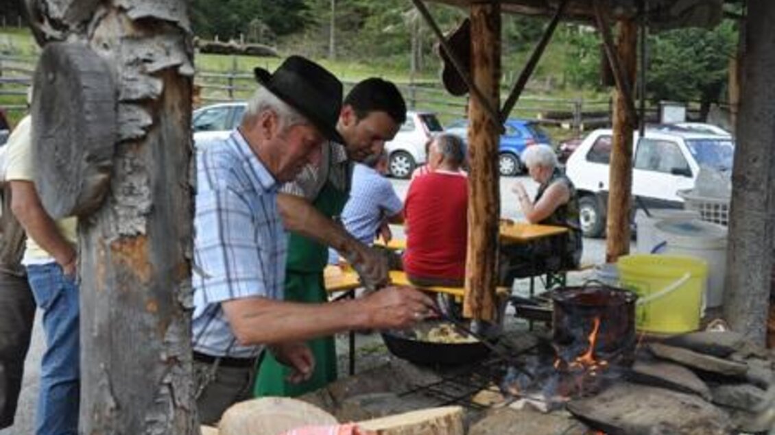 Ein Mann kocht an einem rustikalen Stand im Freien, während ein zweiter Mann ihm hilft. Im Hintergrund sind mehrere Personen an Tischen und parkende Autos zu sehen.