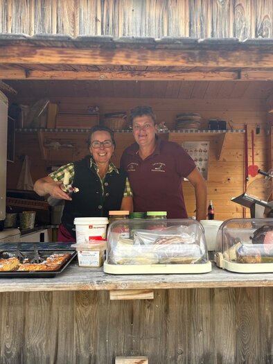 Two friendly men are standing behind a wooden stall and smiling at the camera. Various foods are visible in display cases at the stall.
