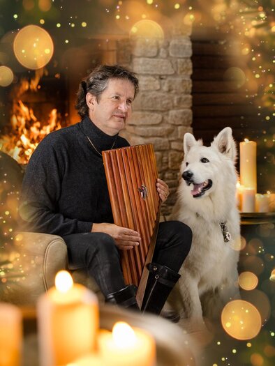 A man is sitting in a cozy room next to a white dog and playing a wooden recorder. In the background, a fireplace is burning and there is candlelight. | © Wolfgang Niegelhell