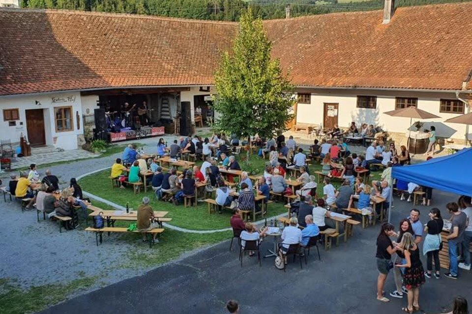 Eine lebhafte Veranstaltung im Freien mit vielen Menschen, die an Tischen sitzen. Im Hintergrund gibt es eine Bühne und eine grüne Umgebung. | © Trummerhof