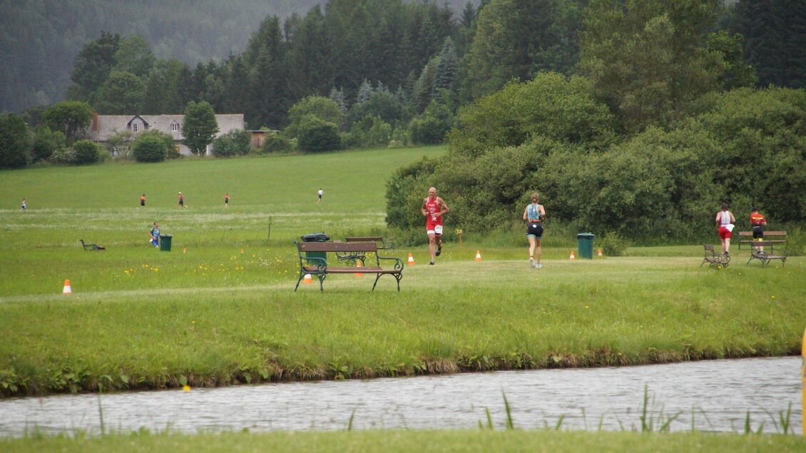 Läufer am Ufer eines ruhigen Gewässers, umgeben von grünen Wiesen und Bäumen. Einige Bänke sind im Hintergrund sichtbar. | © funsports