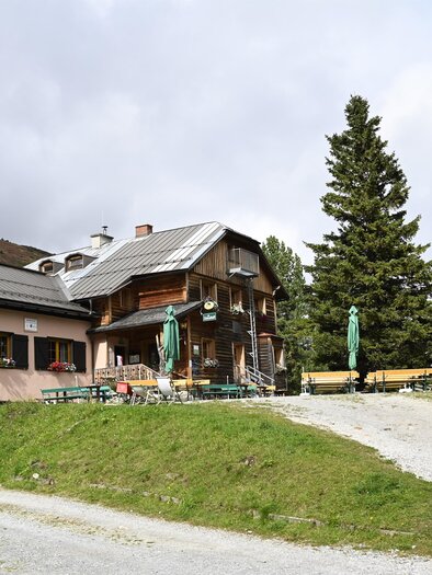 A cozy mountain lodge surrounded by pine trees. The path leads to the terrace with tables and chairs outside. | © Anita Fössl