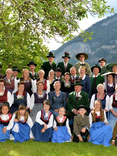 Eine Gruppe von Menschen in traditioneller Tracht steht im Freien. Im Hintergrund sind Bäume und Berge zu sehen.