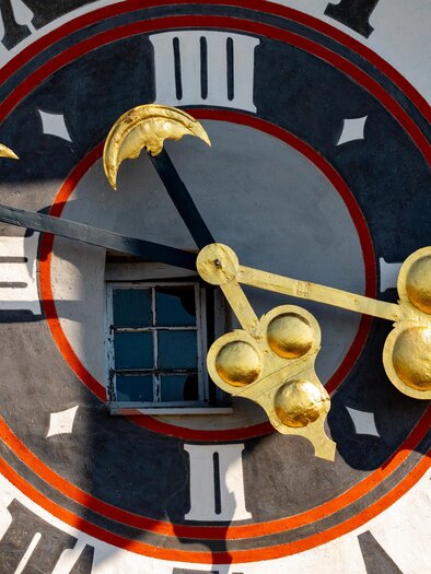 A large, colorful clock with golden hands and Roman numerals. In the background, there is a window. | © Graz Tourismus - Harry Schiffer