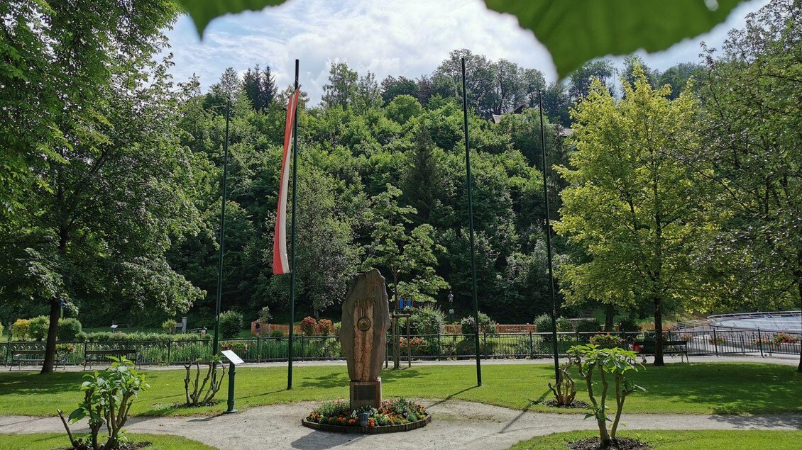<p>Der gepflegte Kurpark in Bad Aussee mit grünen Bäumen und bunten Blumen. Im Mittelpunkt steht der Mittelpunktstein, daneben steht die österreichische Flagge.</ | © TVB Ausseerland - Salzkammergut, www.ausseerland.at