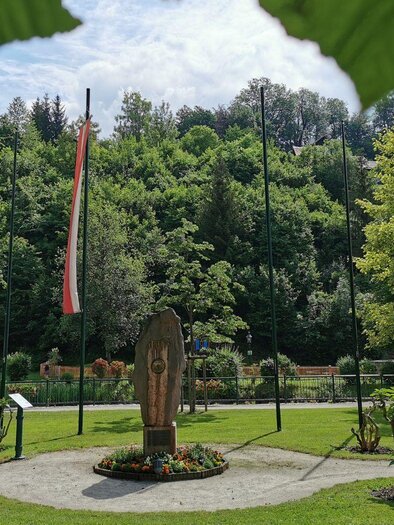 <p>Der gepflegte Kurpark in Bad Aussee mit grünen Bäumen und bunten Blumen. Im Mittelpunkt steht der Mittelpunktstein, daneben steht die österreichische Flagge.</ | © TVB Ausseerland - Salzkammergut, www.ausseerland.at