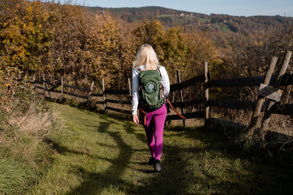 Eine Frau wandert auf einem grünen Weg durch eine herbstliche Landschaft. Im Hintergrund sind Bäume und ein Holzzaun zu sehen. | © Oststeiermark Tourismus