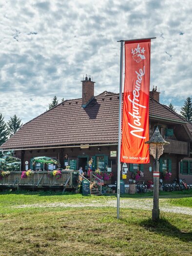 Ein großes Gebäude in der Natur mit einer Terrasse und vielen Pflanzen. Ein Banner mit der Aufschrift "Naturfreunde" ist sichtbar. | © Natufreunde / Doris List-Winder