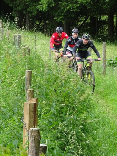 Eine Gruppe von Mountainbikern fährt auf einem schmalen Weg durch eine grüne Wiese. Sträucher und Zäune umrahmen den Pfad, umgeben von Bäumen. | © Eva Reindl