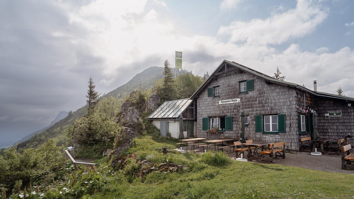 Eine malerische Berghütte umgeben von grünen Wiesen und Bäumen. Im Hintergrund sind hohe Berge und ein bewölkter Himmel zu sehen. | © TV Gesäuse