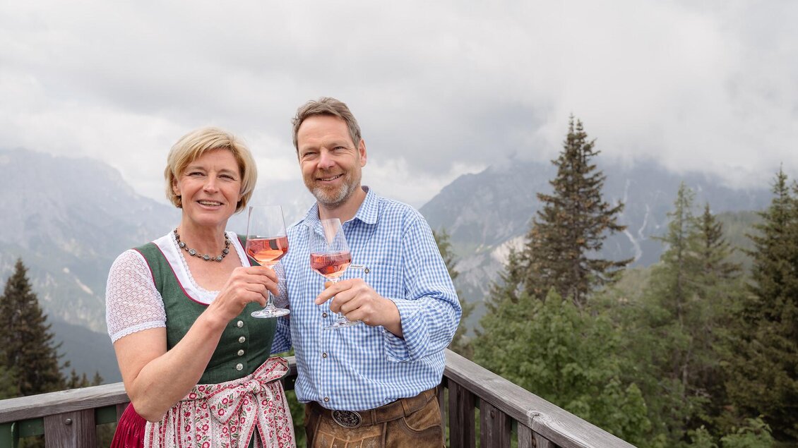 Ein Paar in traditioneller Kleidung steht auf einer Veranda mit Blick auf die Berge. Sie halten Gläser mit Getränken und lächeln. | © Tourismusverband Gesäuse