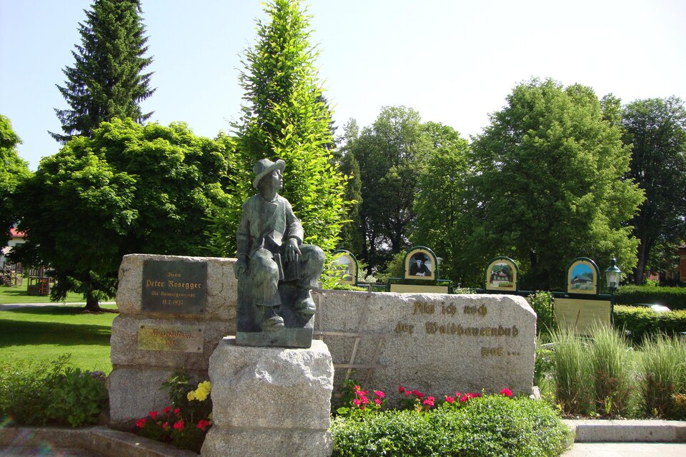 Eine Skulptur eines Kindes mit einem Hund steht auf einem Steinblock in einem grünen Park. Im Hintergrund sind Bäume und bunte Blumen zu sehen. | © Gemeinde Krieglach