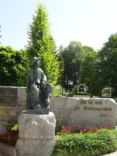 Eine Skulptur eines Kindes mit einem Hund steht auf einem Steinblock in einem grünen Park. Im Hintergrund sind Bäume und bunte Blumen zu sehen. | © Gemeinde Krieglach