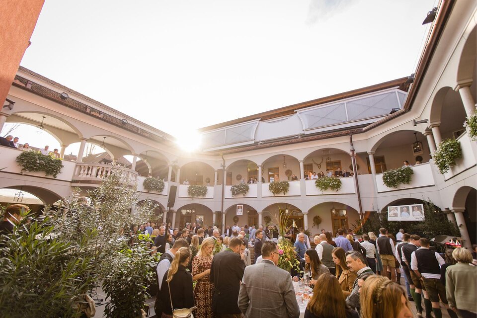 Eine lebhafte Veranstaltung in einem eleganten Innenhof mit vielen Gästen. Die Sonne strahlt durch die Bögen und sorgt für eine freundliche Atmosphäre.