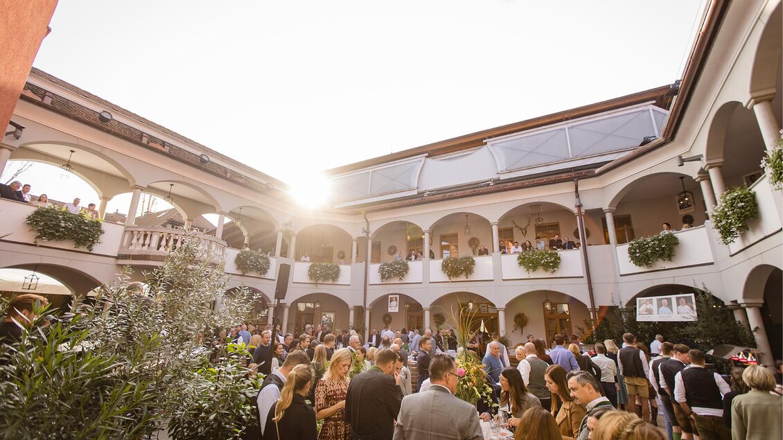 Eine lebhafte Veranstaltung in einem eleganten Innenhof mit vielen Gästen. Die Sonne strahlt durch die Bögen und sorgt für eine freundliche Atmosphäre.