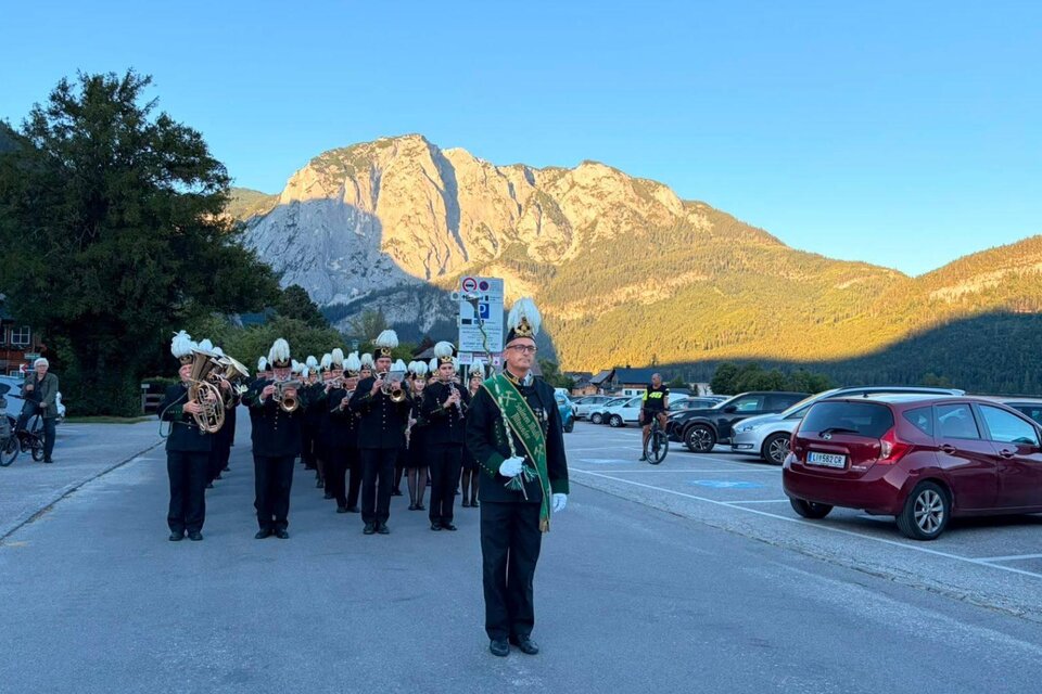 Eine Musikparade mit mehreren Musikern in schwarzen Anzügen und weißen Hüten findet vor einer beeindruckenden Berglandschaft statt. Im Vordergrund steht ein Mann mit einer Schärpe und einem Taktstock. | © Salinenmusik Altaussee_Theresa Rastl_Robert Leu