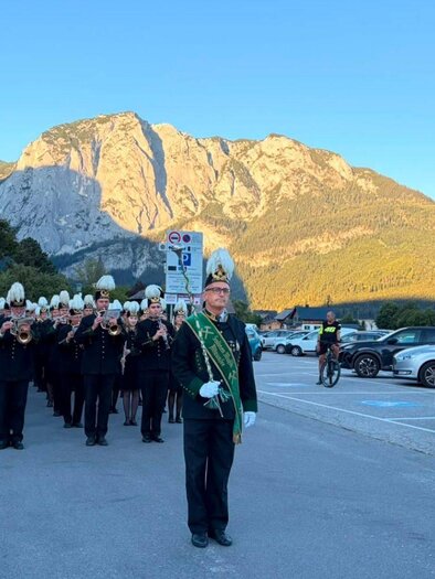 Eine Musikparade mit mehreren Musikern in schwarzen Anzügen und weißen Hüten findet vor einer beeindruckenden Berglandschaft statt. Im Vordergrund steht ein Mann mit einer Schärpe und einem Taktstock. | © Salinenmusik Altaussee_Theresa Rastl_Robert Leu