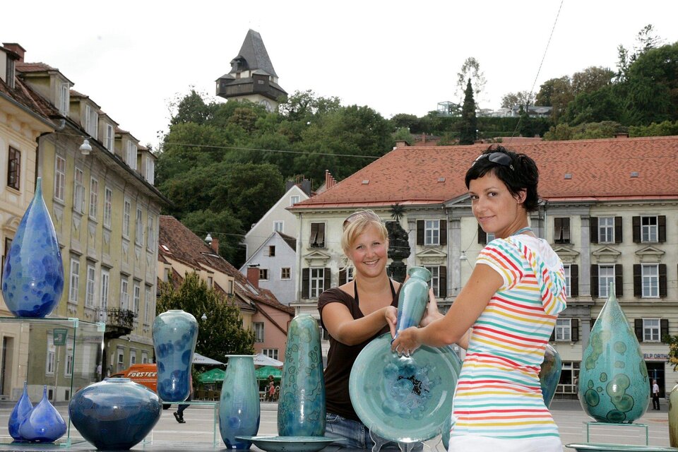 Zwei Frauen stehen auf einem Platz und präsentieren farbige Glasobjekte. Im Hintergrund sind historische Gebäude und ein Hügel mit einem Aussichtsturm zu sehen. | © Graz Tourismus - Foto Fischer