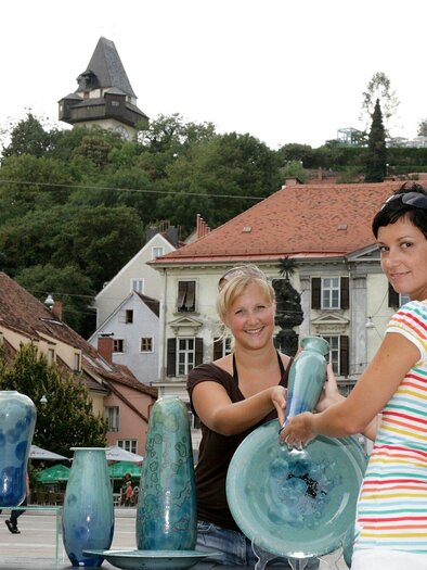 Zwei Frauen stehen auf einem Platz und präsentieren farbige Glasobjekte. Im Hintergrund sind historische Gebäude und ein Hügel mit einem Aussichtsturm zu sehen. | © Graz Tourismus - Foto Fischer