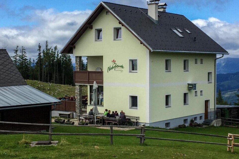 Ein modernes, gelbes Haus inmitten einer malerischen Berglandschaft. Draußen sitzen Menschen in einem einladenden Außenbereich. | © Walter Riesenmann