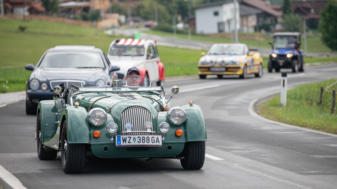 Ein klassischer Morgan-Sportwagen fährt auf einer Landstraße, umgeben von anderen Autos. Im Hintergrund sieht man ländliche Gebäude und eine grüne Landschaft. | © TV Hochsteiermark, E. Paller