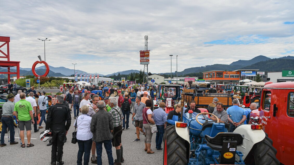 Eine große Menschenmenge versammelt sich auf einem Parkplatz mit Traktoren und alten Fahrzeugen. Im Hintergrund sind Geschäfte und Berge sichtbar. | © E.Paller