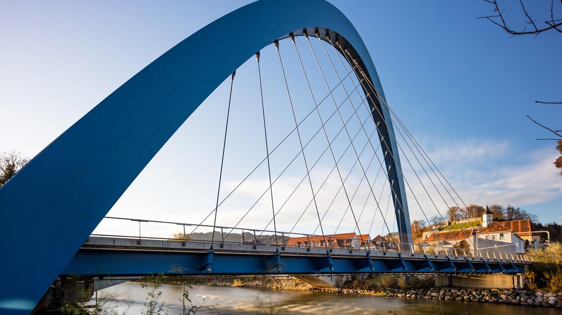 Eine moderne blaue Brücke mit einem eleganten Bogen. Im Hintergrund fließt ein Fluss und es ist ein klarer Himmel sichtbar. | © Stadt_Bruck, M.Meieregger