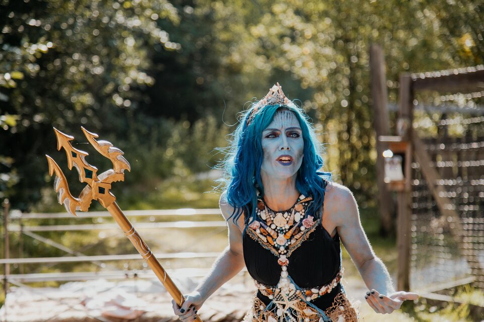 Eine Person mit blauen Haaren steht im Freien und trägt ein auffälliges Kostüm mit einer Trident. Sie sieht entschlossen aus und ist von grüner Natur umgeben. | © Naturpark Mürzer Oberland,  Nici Seiser