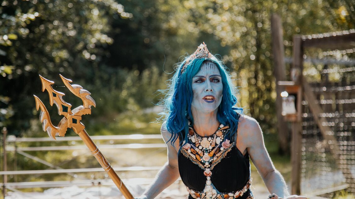 Eine Person mit blauen Haaren steht im Freien und trägt ein auffälliges Kostüm mit einer Trident. Sie sieht entschlossen aus und ist von grüner Natur umgeben. | © Naturpark Mürzer Oberland,  Nici Seiser