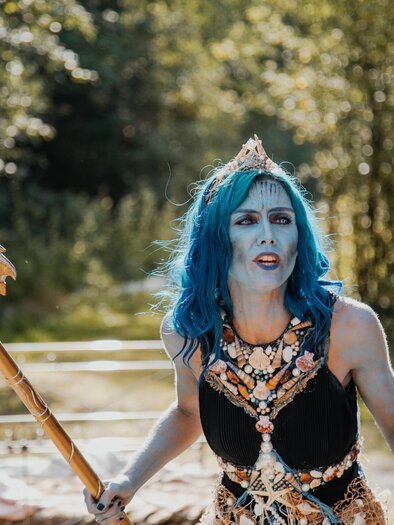 Eine Person mit blauen Haaren steht im Freien und trägt ein auffälliges Kostüm mit einer Trident. Sie sieht entschlossen aus und ist von grüner Natur umgeben. | © Naturpark Mürzer Oberland,  Nici Seiser