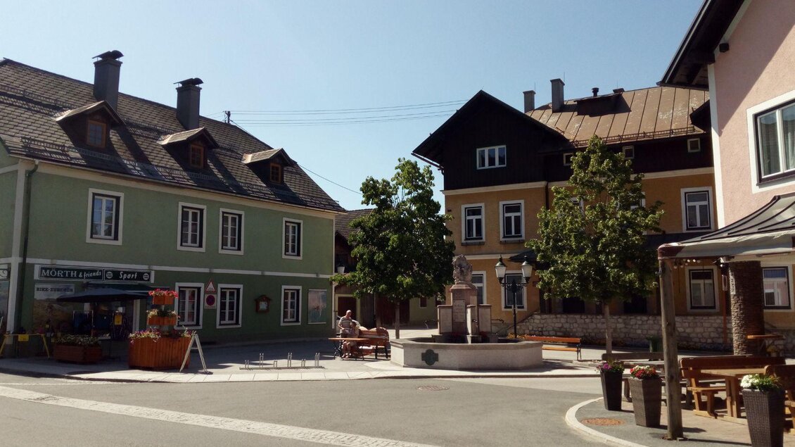 Blick auf den Dorfplatz mit dem Sportgeschäft Mörth im Hintergrund. Gut erkennbar sind der Brunnen sowie die Sitzbänke und Bäume, die den Platz umgeben. Der strahlend blaue Himmel sorgt für eine freundliche und einladende Atmosphäre. | © Johanna Provatopoulos