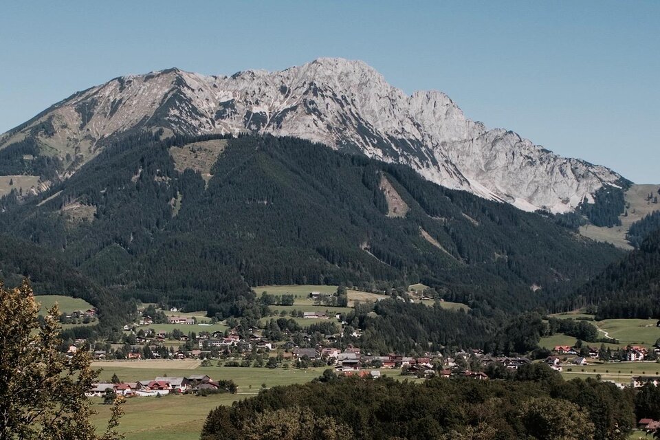 Eine beeindruckende Berglandschaft mit hohen Bergen und saftig grünen Tälern. Im Vordergrund ist ein kleines Dorf eingebettet in die Landschaft. | © Kren Ernst