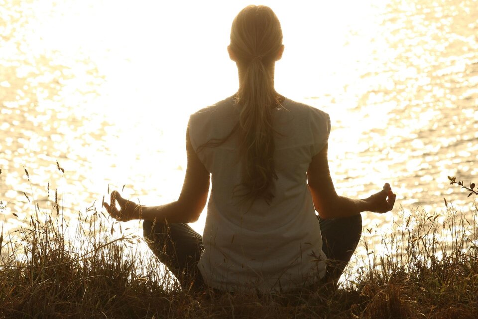 Eine Person sitzt in meditativer Haltung am Ufer eines Gewässers. Die Sonne reflektiert sich auf der Wasseroberfläche und erzeugt eine friedliche Atmosphäre. | © Pixabay