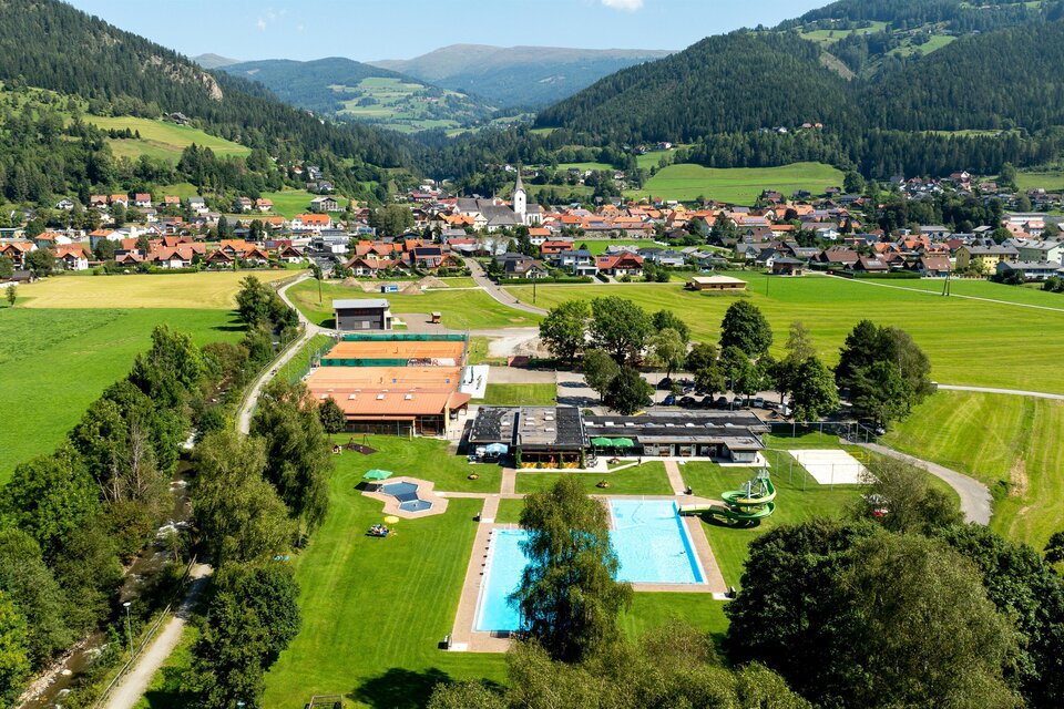Eine malerische Landschaft mit einem Hotelkomplex und einem Poolbereich. Im Hintergrund sind grüne Hügel und ein kleines Dorf zu sehen. | © Tv Murau