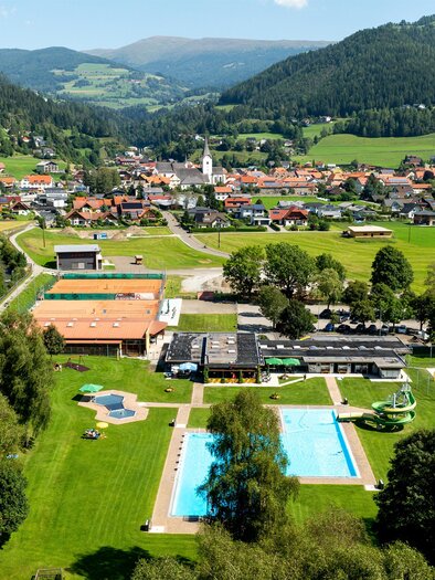 A picturesque landscape with a hotel complex and a pool area. In the background, green hills and a small village can be seen. | © Tv Murau