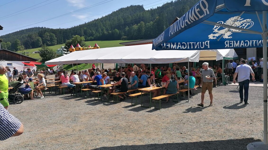Eine lebhafte Veranstaltung im Freien mit vielen Menschen, die an Tischen sitzen. Im Hintergrund sind grüne Hügel und ein klarer blauer Himmel zu sehen. | © Apfelmaier