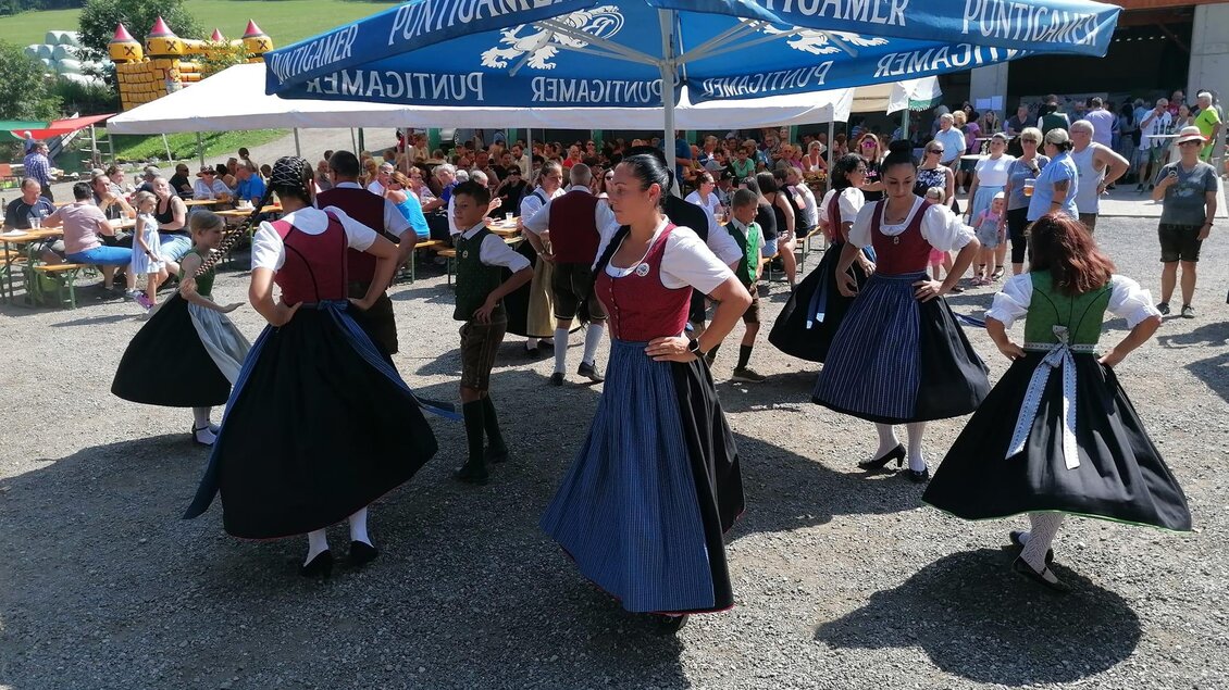 Eine folkloristische Tanzaufführung findet unter einem großen Zeltdach statt. Die Tänzerinnen tragen traditionelle Trachten und es sind viele Zuschauer im Hintergrund zu sehen. | © Apfelmaier