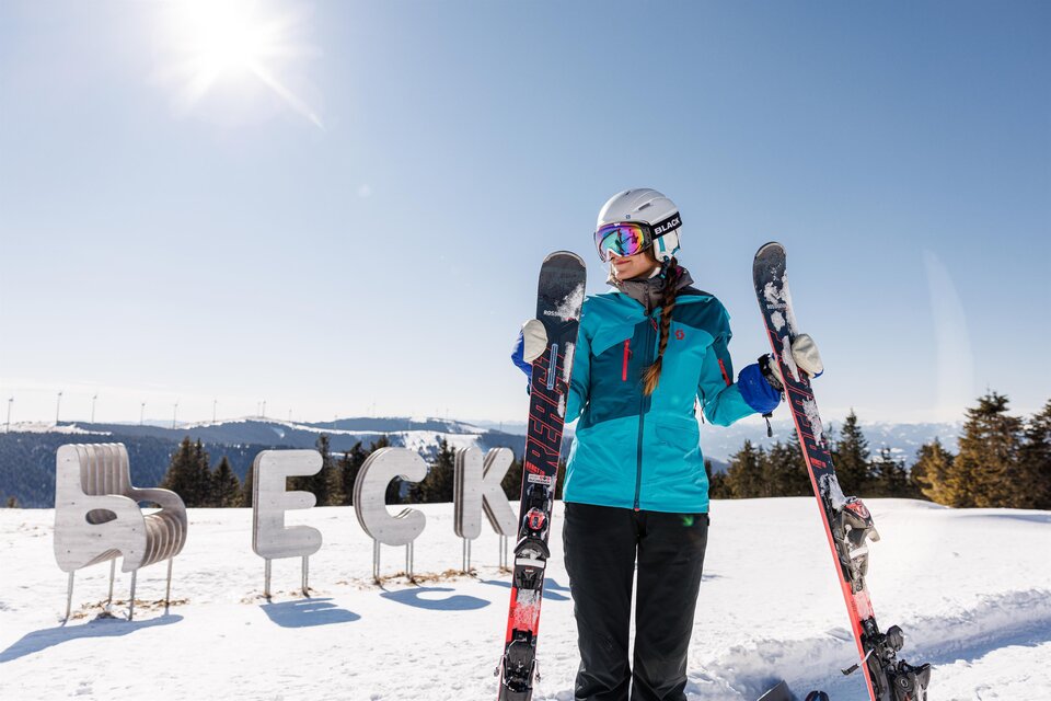 Eine Skifahrerin steht im Schnee und hält ihre Skier in den Händen. Im Hintergrund sind schneebedeckte Berge und der Schriftzug "ECK" zu sehen. | © nicoleseiser
