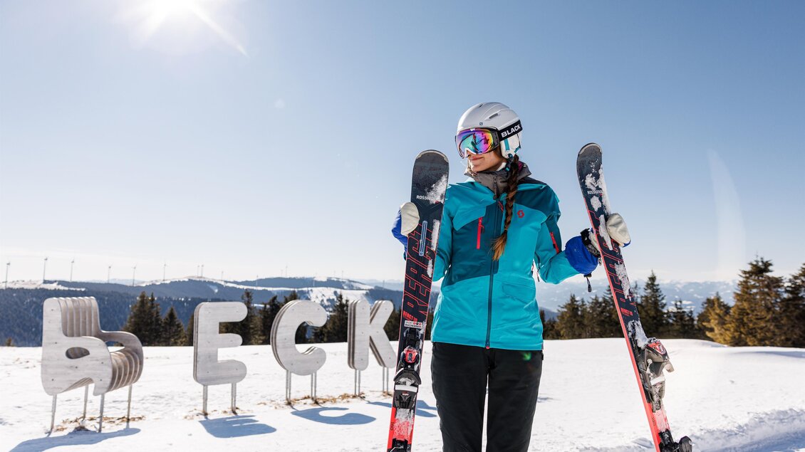 Eine Skifahrerin steht im Schnee und hält ihre Skier in den Händen. Im Hintergrund sind schneebedeckte Berge und der Schriftzug "ECK" zu sehen. | © nicoleseiser