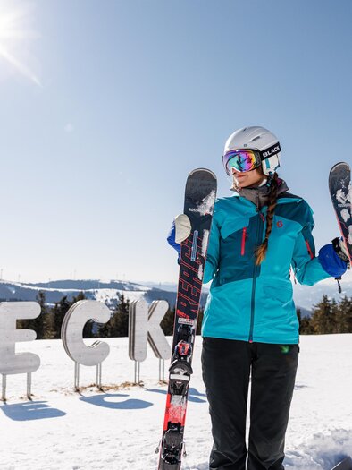 Eine Skifahrerin steht im Schnee und hält ihre Skier in den Händen. Im Hintergrund sind schneebedeckte Berge und der Schriftzug "ECK" zu sehen. | © nicoleseiser
