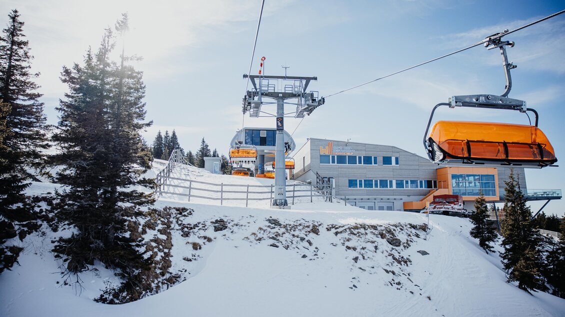 Eine Skiliftanlage mit orangefarbenen Sesseln in einer verschneiten Landschaft. Im Hintergrund sieht man ein modernes Gebäude und Tannenbäume. | © nicoleseiser