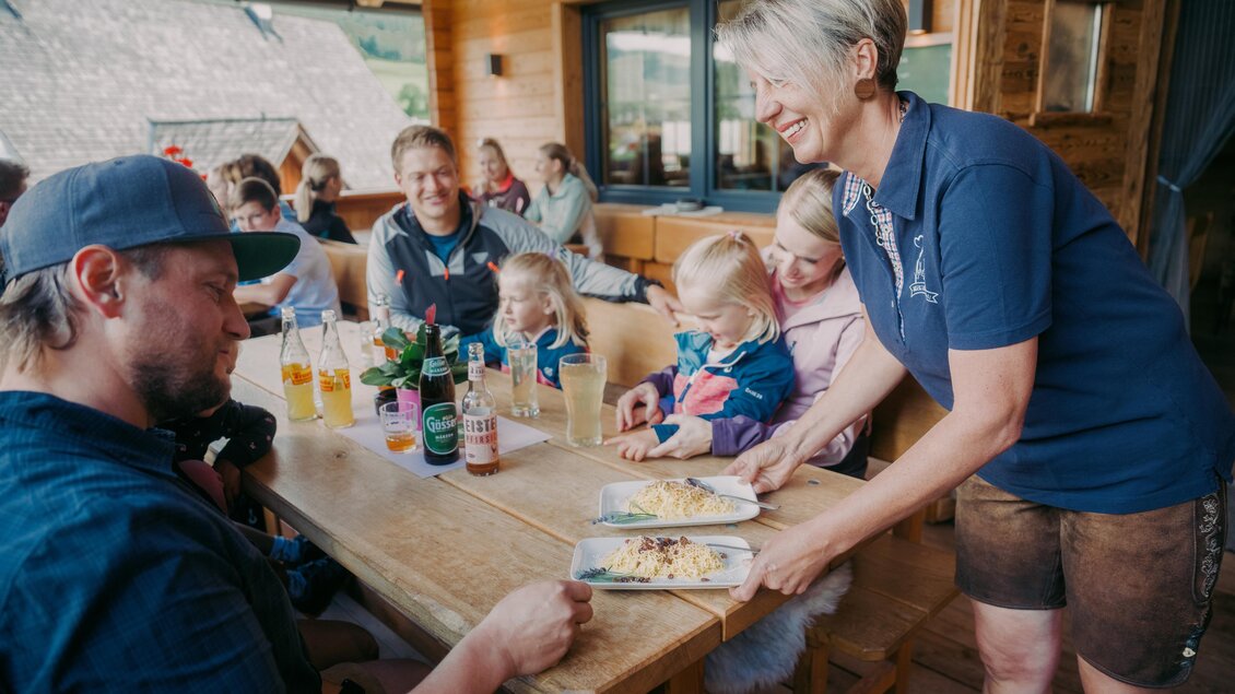 Eine fröhliche Gruppe von Menschen sitzt an einem Holztisch und genießt die gemeinsame Zeit. Eine Kellnerin serviert Essen an einen Gast. | © Roman Königshofer