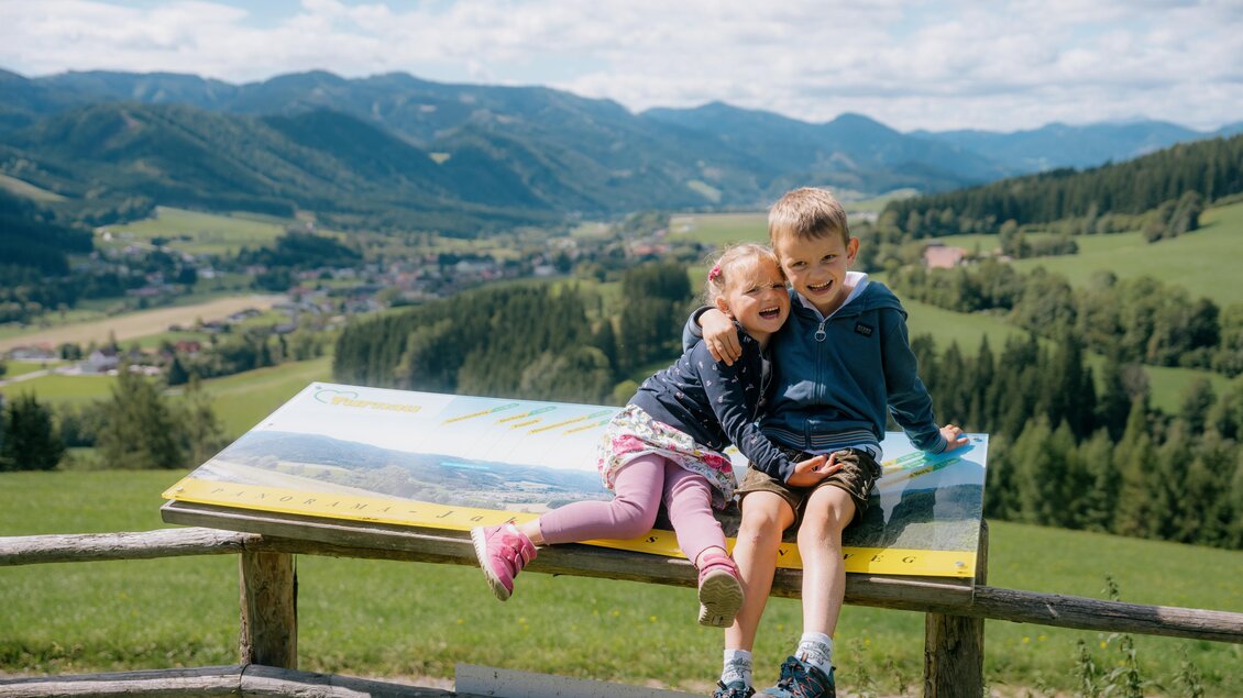 Zwei Kinder sitzen auf einer Bank mit Blick auf eine grüne Landschaft und Berge im Hintergrund. Der Himmel ist klar und blau, und die Kinder lächeln glücklich. | © Roman Königshofer